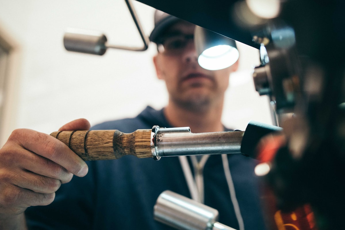 Nahaufnahme einer Hand am Holzgriff einer Metallmaschine, im Hintergrund eine Person mit Kapuzenpullover bei der Bedienung in einer Werkstatt
